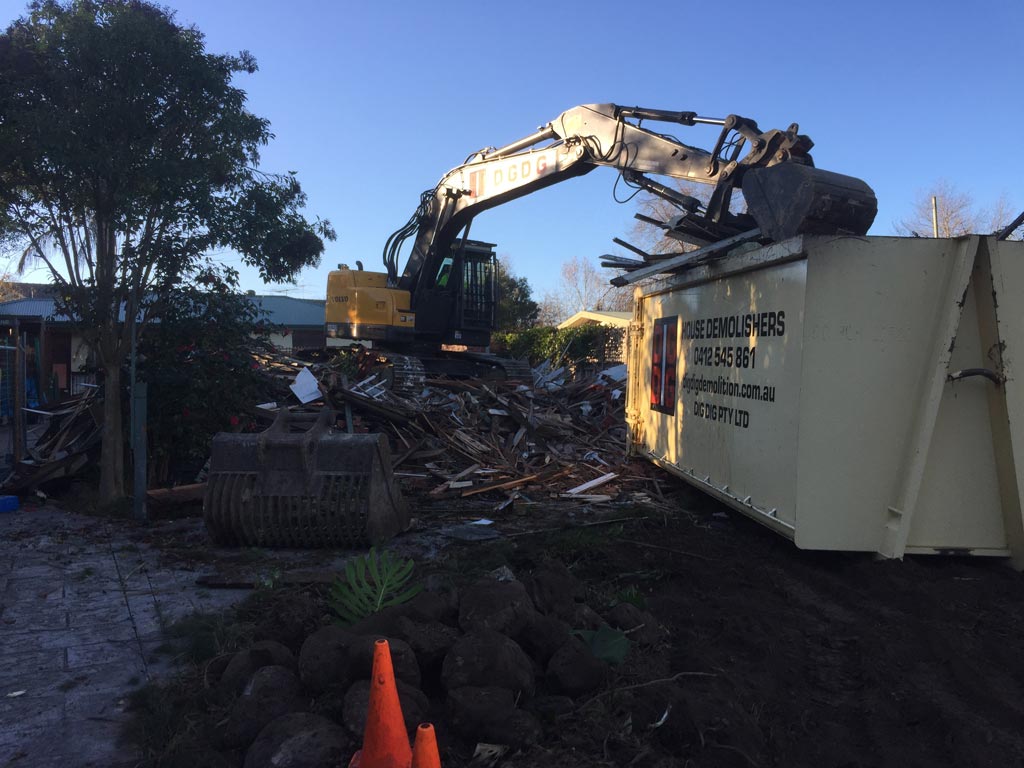 Excavator loading timber debris into a DigDig Demolition skip bin at a house demolition site