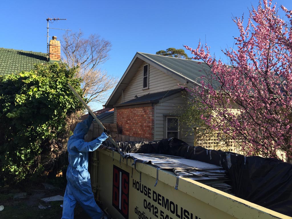 Worker in protective coveralls loading roof sheeting into a covered DigDig Demolition skip bin