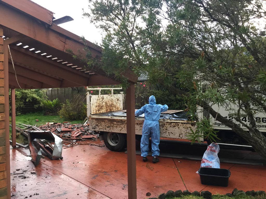Worker in protective coveralls loading material onto a DigDig Demolition truck under a carport